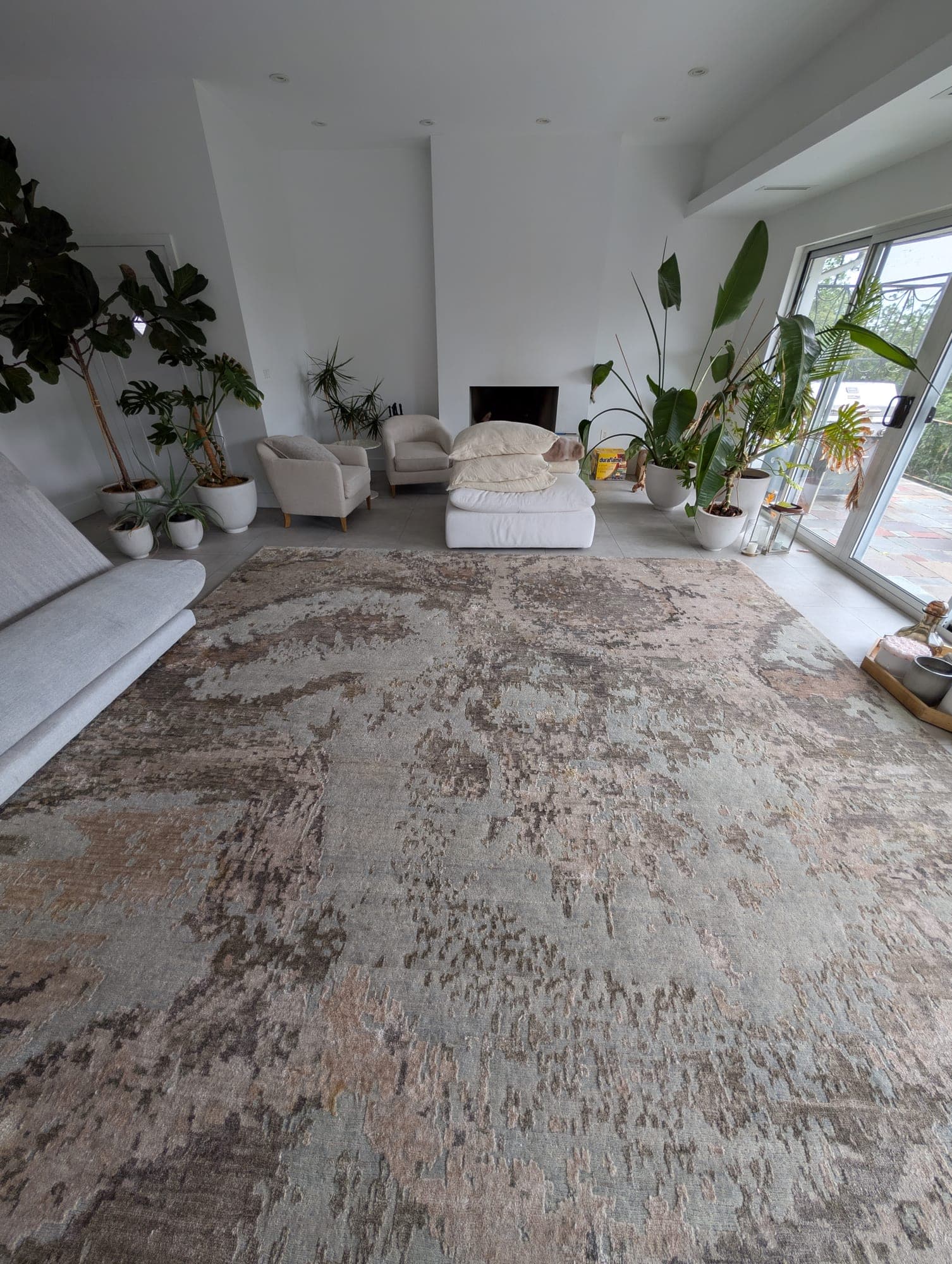Large hand-knotted abstract gray and beige area rug returned to a modern living room with tall plants and fireplace