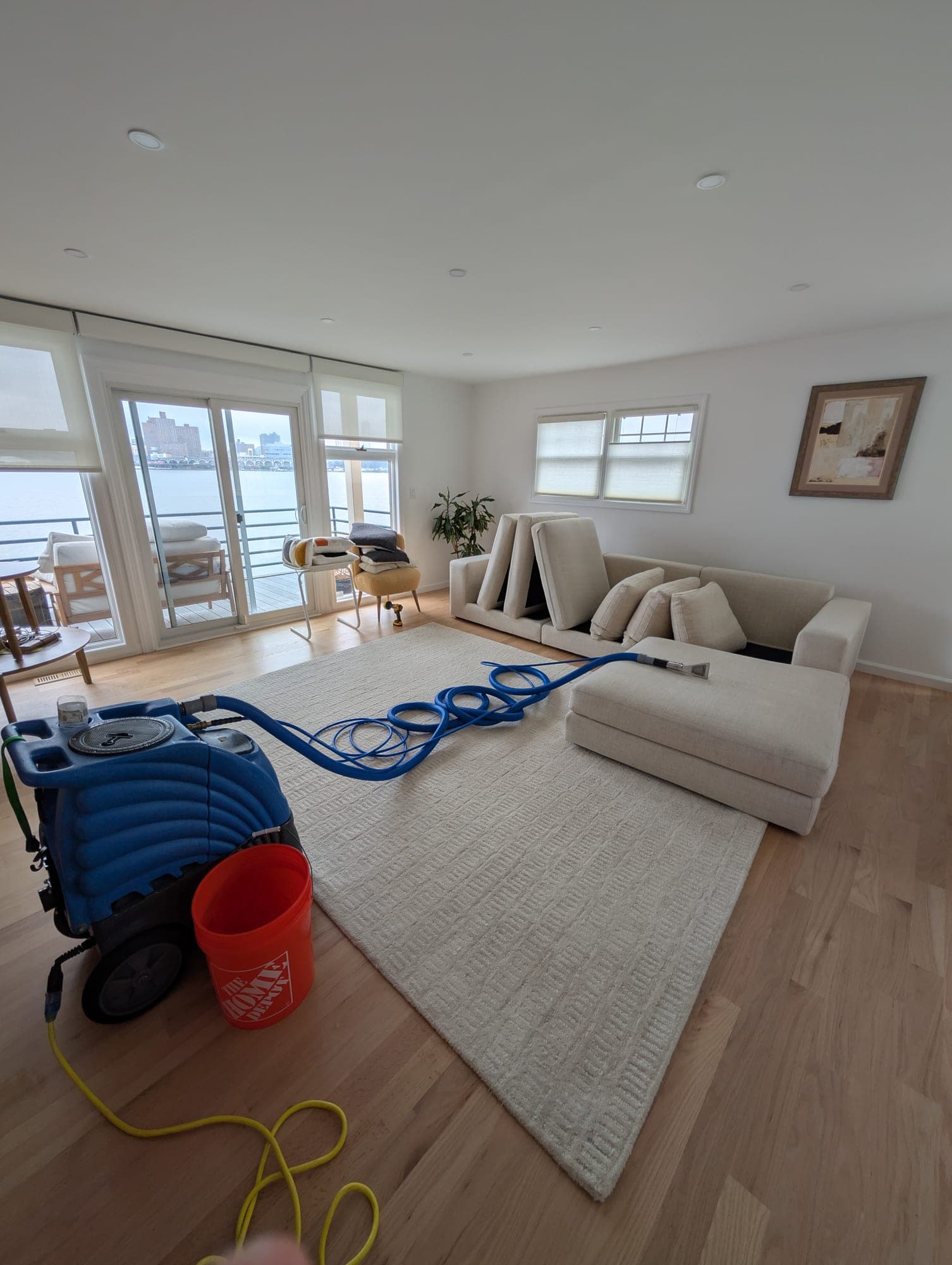 Real Zep job: bright NJ waterfront living room with a freshly cleaned cream wool area rug, hot-water extraction machine on the hardwood floor, and a water view through floor-to-ceiling windows